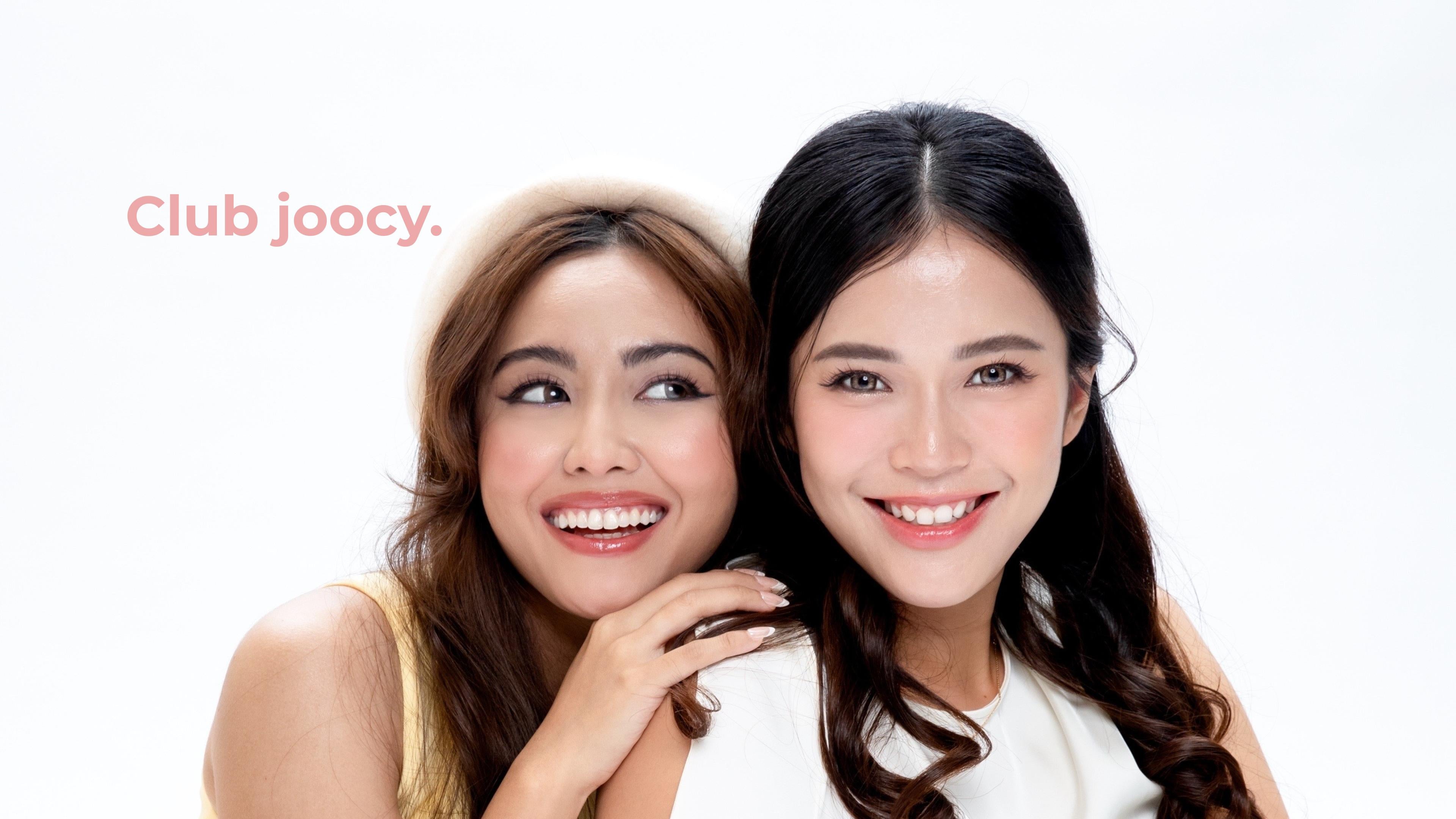 Club joocy. Two models, one tan-skinned Malay and one fair-warm Chinese, smiling happily while wearing coordinating yellow and white outfits. Both are wearing joocy cosmetic contact lenses, demonstrating natural blending on dark eyes and natural glow across diverse Malaysian skin tones.