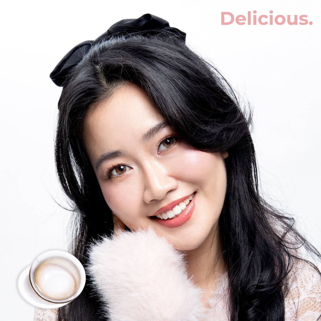 Product page image of a smiling fair-to-medium skin model wearing joocy Korean design cosmetic lenses in shade Delicious, showcasing a hyper-realistic sepia brown color payoff. Includes a macro swatch graphic showing the glassy dimension lens pattern.