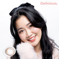 Product page image of a smiling fair-to-medium skin model wearing joocy Korean design cosmetic lenses in shade Delicious, showcasing a hyper-realistic sepia brown color payoff. Includes a macro swatch graphic showing the glassy dimension lens pattern.