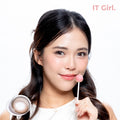 Product page image of a model with a fair-warm skin tone holding a pink lollipop, wearing joocy grey-brown Korean design contact lenses in shade IT Girl. Includes a macro swatch graphic of the single lens showing its deep cocoa and iced-grey doll-like pattern.