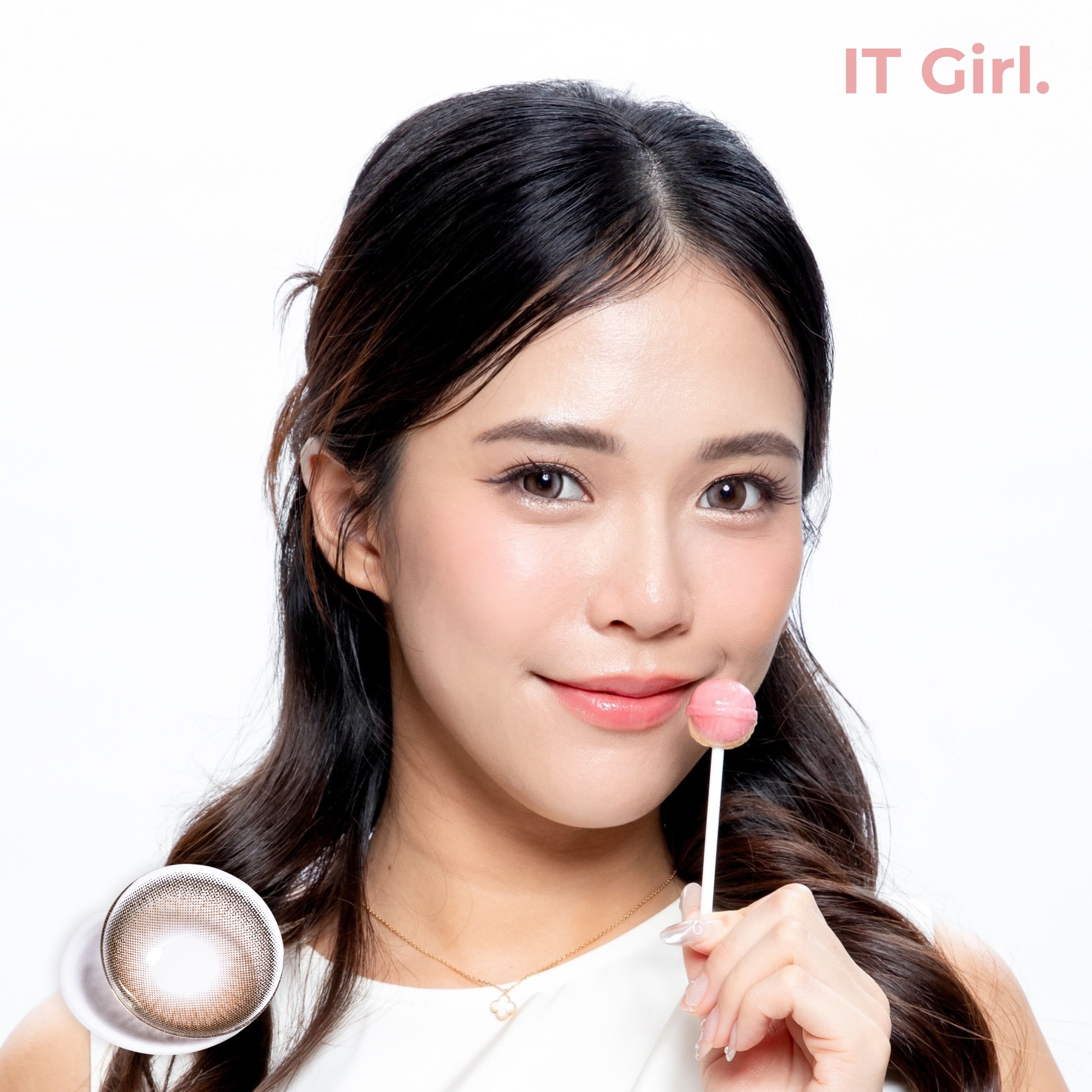 Product page image of a model with a fair-warm skin tone holding a pink lollipop, wearing joocy grey-brown Korean design contact lenses in shade IT Girl. Includes a macro swatch graphic of the single lens showing its deep cocoa and iced-grey doll-like pattern.